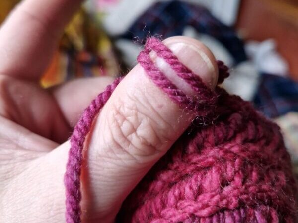 A close-up of a white man's thumb with yarn looped around it, working on a length of pink-purple nåbinding